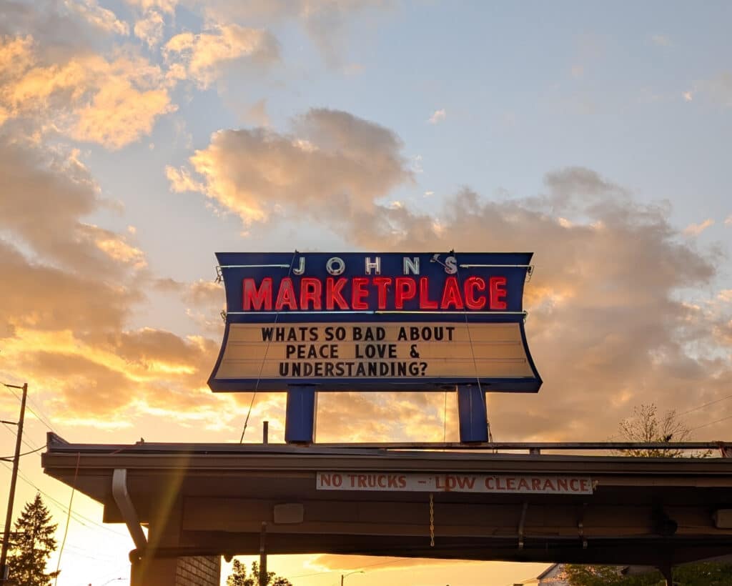 A neon sign reading “JOHN’S MARKETPLACE” with a marquee below that says, “WHATS SO BAD ABOUT PEACE LOVE & UNDERSTANDING?” against a golden sky.
