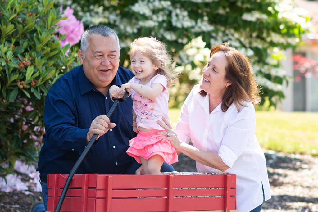 Photo of grandparents playing with their grandchild that is standing and laughing in a red wagon