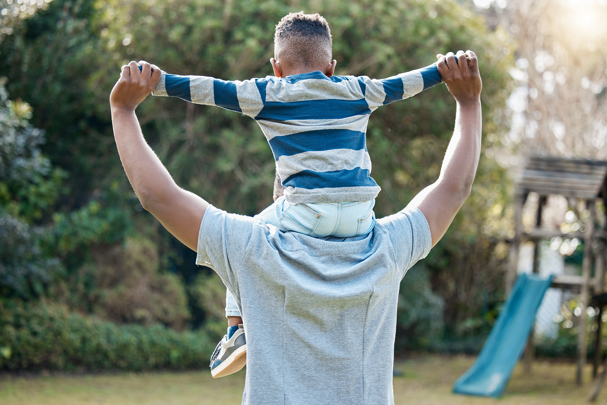 A child sitting atop their parents shoulders, both are holding hands outwards and being playful.