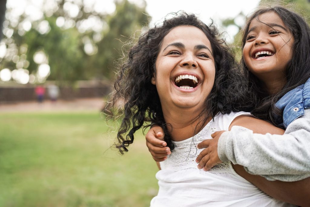 A daughter riding on her mother's shoulder outside, both are laughing playfully.