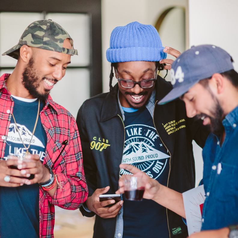 A group of men smiling and laughing at a community event, wearing Live Like the Mountain is Out shirts