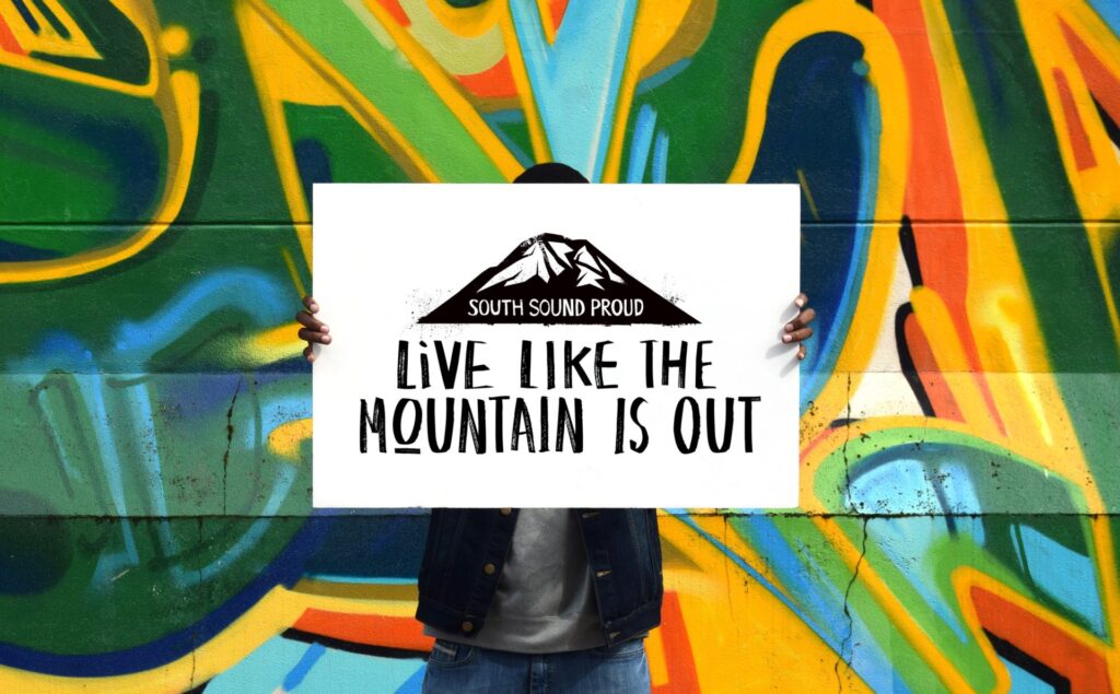 A person stands against a colorful wall mural holding a sign reading "South Sound Proud: Live Like the Mountain is Out"