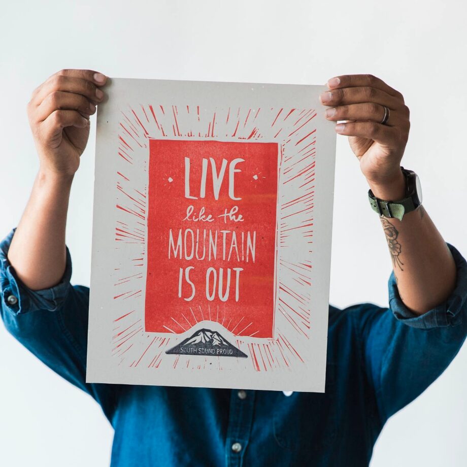 A person holds up a letterpress Live Like the Mountain is Out poster