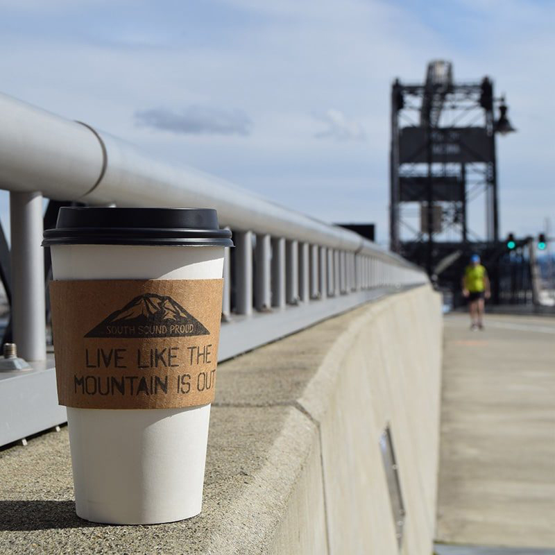 A coffee cup wearing a sleeve stamped with Live Like the Mountain is Out sits along a railing in front of a draw bridge