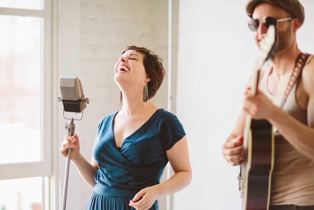 A woman joyously sings into an old-fashioned microphone next to a man playing acoustic guitar at a community event