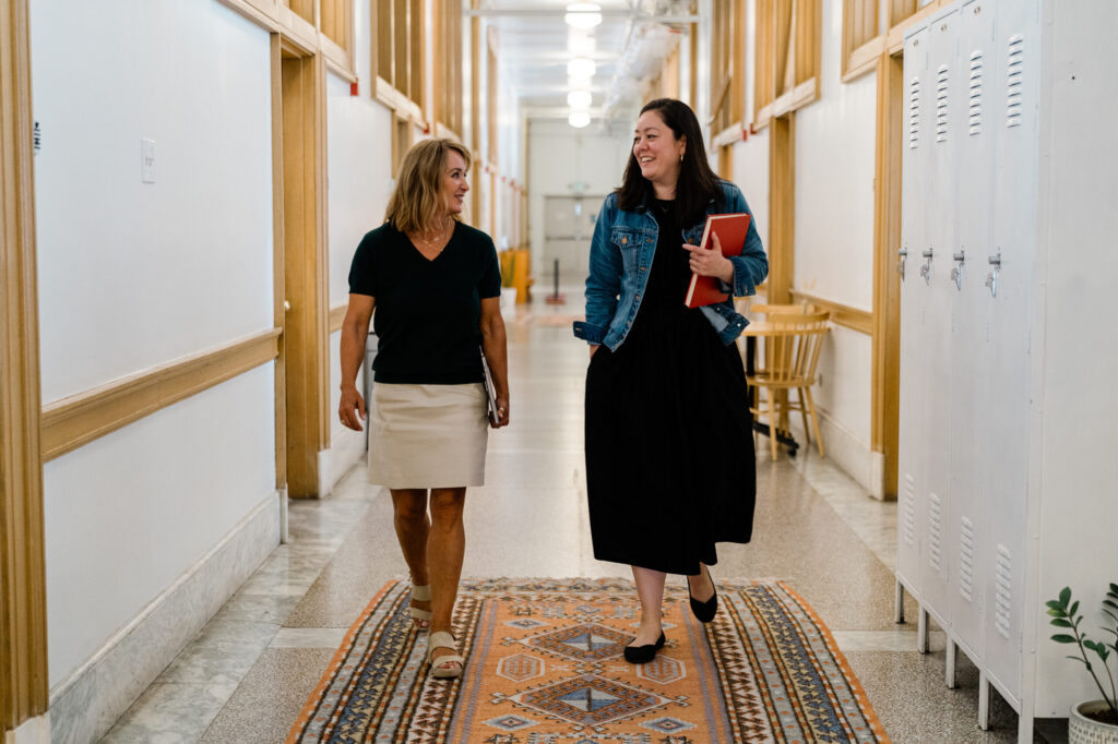 Two women walk down an office hallway, one holding a red notebook