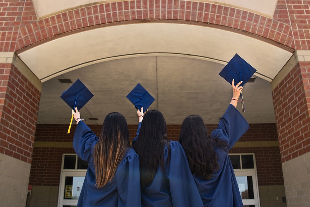 Three graduates stand in front of a brick building holding their caps in the air